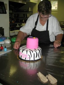 food: a woman in a white top and black apron decorating a cake