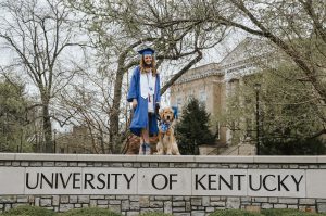 a golden retriever in a graduation cap and a woman in a cap and gown standing on a wall that says university of kentucky